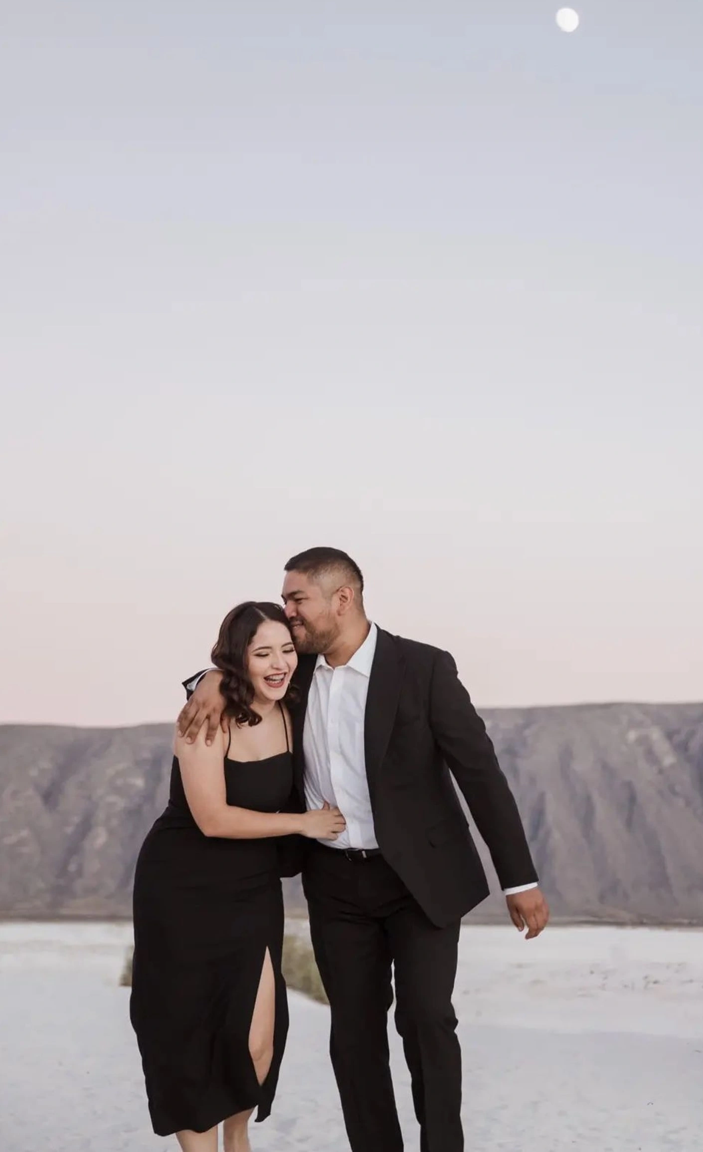 Una pareja sonriendo vestida de negro; él besa la sien de ella frente a un paisaje desértico con montañas, bajo un cielo claro donde se ve la luna.