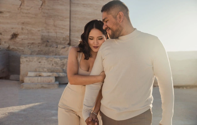 Pareja caminando tomada de la mano bajo la luz del sol, compartiendo una mirada cómplice en un escenario natural.